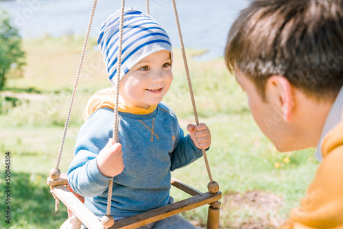 Stylish young father swings his happy three-year-old son dressed in yellow blue clothes on a wooden swing on a sunny day in the countryside. The concept of child care, safety for children. photo