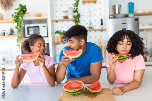 Portrait of cheerful young family taking a bite of a watermelon. African American family standing together at a kitchen eating watermelon.