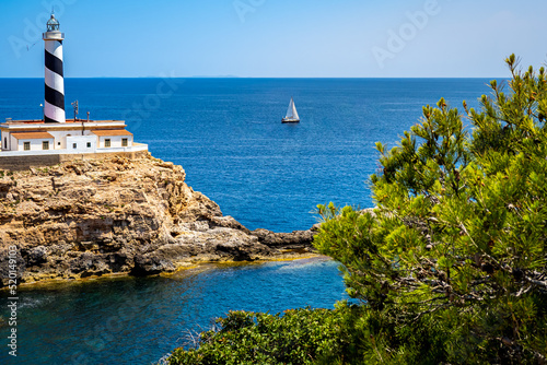 Tableau sur toile Panorama view of the Mallorca Cala Figuera lighthouse on a peninsula with an idyllic cove and a sailboat in front of the mediterranean sea and islands Illa des Conills and Cabrera on the horizon