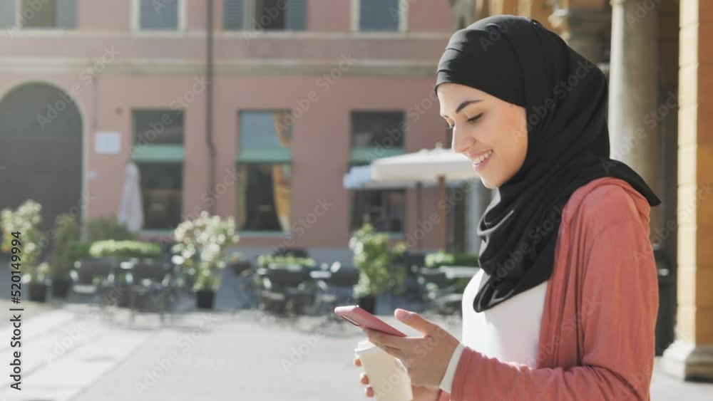 muslim female using smart phone mobile walks in european city square,pleased arab girl wearing hijab uses smartphone while walking tracking shot steadicam slow motion side view