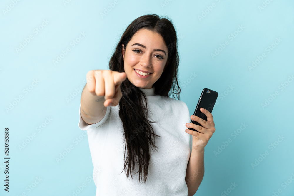 Young woman using mobile phone isolated on blue background points finger at you with a confident expression Young woman using mobile phone isolated on blue background points finger at you with a confident expression