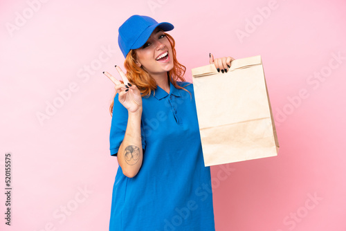 Young caucasian woman taking a bag of takeaway food isolated on pink background smiling and showing victory sign