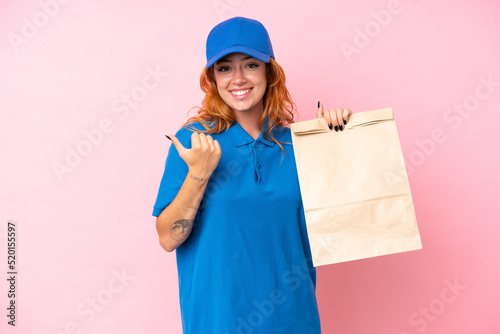Young caucasian woman taking a bag of takeaway food isolated on pink background pointing to the side to present a product