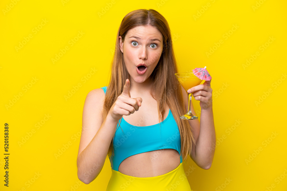 Young caucasian woman holding a cocktail isolated on yellow background surprised and pointing front Young caucasian woman holding a cocktail isolated on yellow background surprised and pointing front