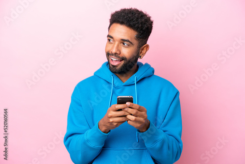 Young Brazilian man isolated on pink background using mobile phone and looking up