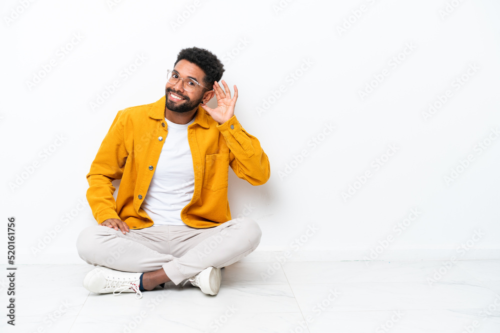 Young Brazilian man sitting on the floor isolated on white background listening to something by putting hand on the ear Young Brazilian man sitting on the floor isolated on white background listening to something by putting hand on the ear