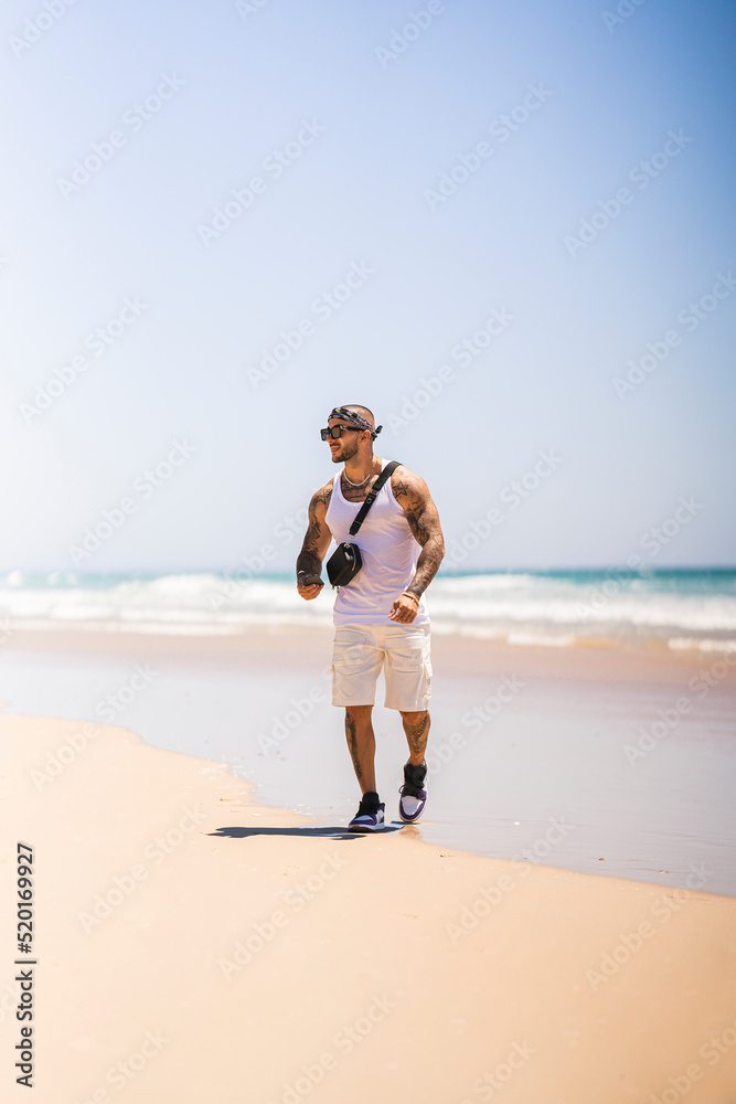 Chico joven tatuado y musculado en playa preciosa en día soleado con camiseta de tirantas blanca y pañuelo en la cabeza Chico joven tatuado y musculado en playa preciosa en día soleado con camiseta de tirantas blanca y pañuelo en la cabeza