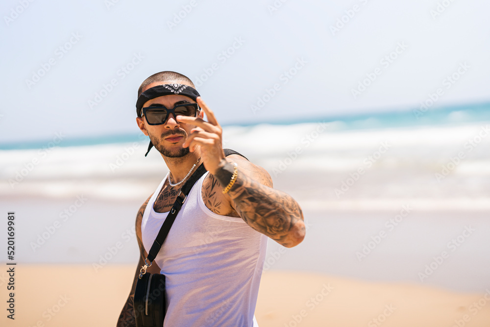 Chico joven tatuado musculoso en la playa con camiseta de tirantas blanca y pañuelo en la cabeza