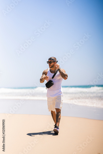 Chico joven tatuado y musculado en playa preciosa en día soleado con camiseta de tirantas blanca y pañuelo en la cabeza