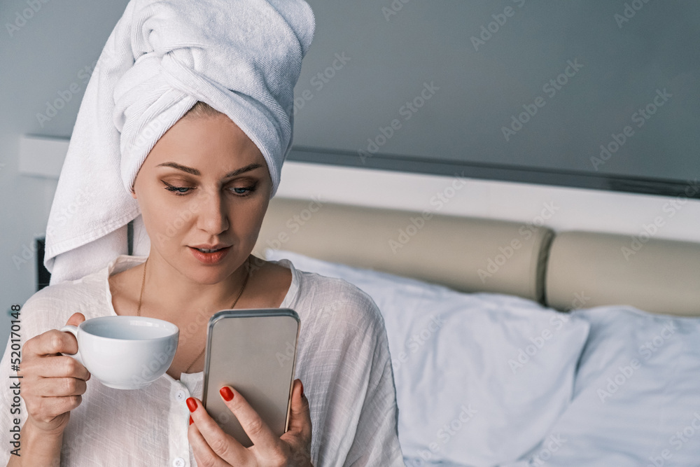 Young attractive millennial woman uses her phone reading the news, scrolling a social media feed sitting with a white poloton on her head in bedroom bed with a cup in her hands. Concept of the morning