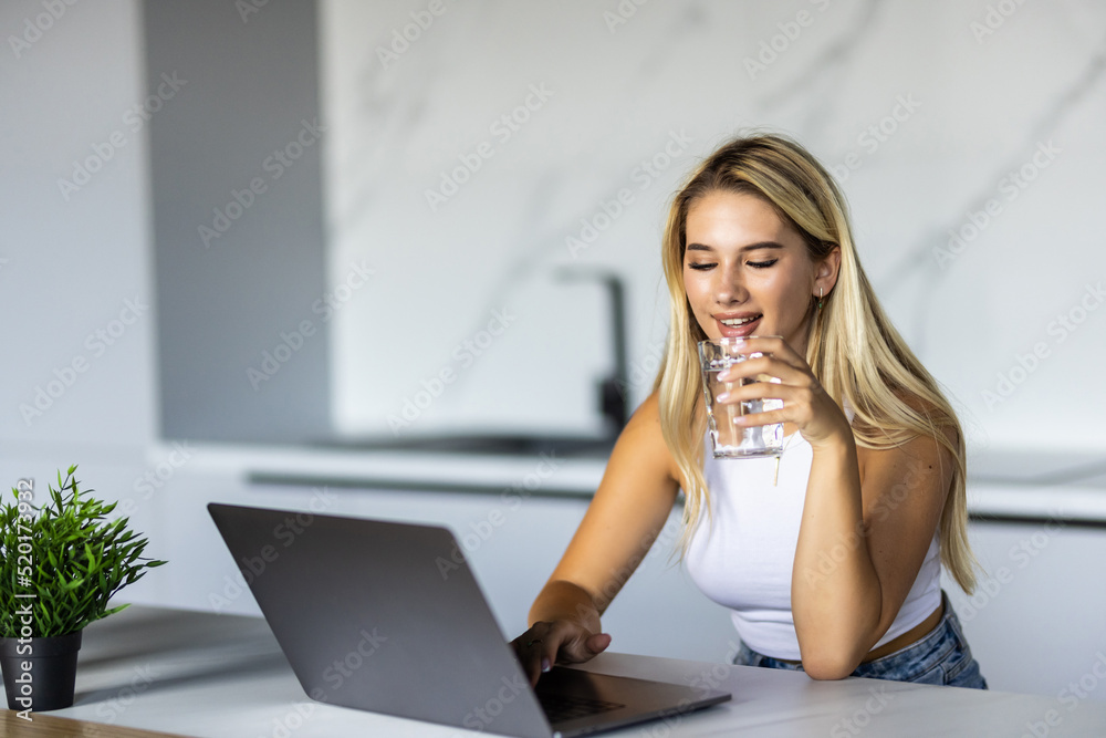 Fototapeta premium Young woman sitting in kitchen table, looking at laptop computer screen. Female entrepreneur working sitting at a desk typing on her laptop in a home office