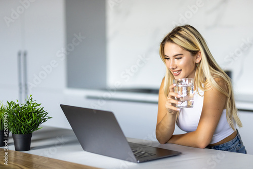 Shot of beautiful woman working with laptop while drinking glass of water on desk in office at home. Refreshment, hydration and Healthy habit concept.