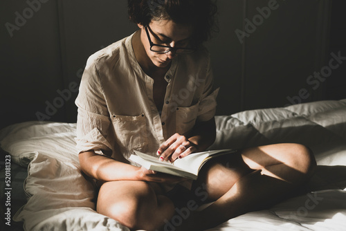 Canvas Print Young woman reading in her room with the light from the window sitting on the bed