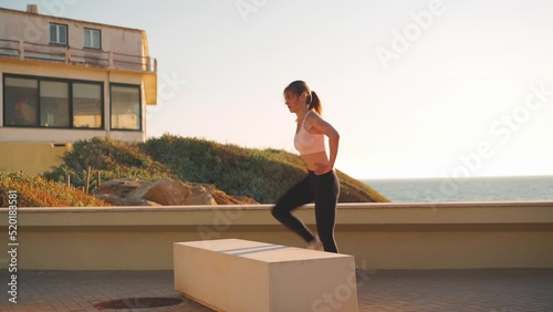 Slim female fitness step-up exercise jump box, strengthening and toning quadriceps and hamstrings while practicing cross training at outdoor gym. Athletic caucasian female workout sea view background