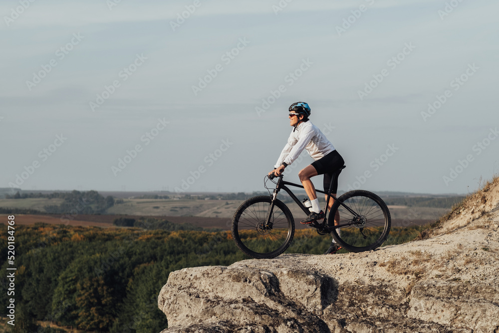 Equipped Professional Male Cyclist Standing with Mountain Bike on Edge of the Rock and Enjoys Panoramic View Outdoors