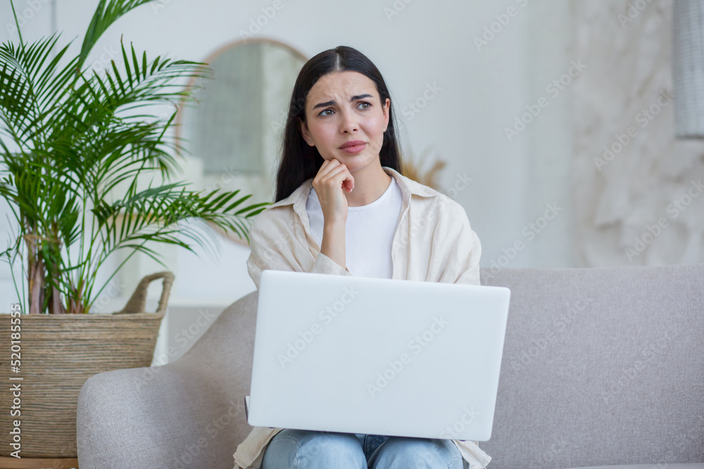 Sad and upset woman in despair reading bad news from laptop, brunette in glasses sitting on sofa at home Sad and upset woman in despair reading bad news from laptop, brunette in glasses sitting on sofa at home