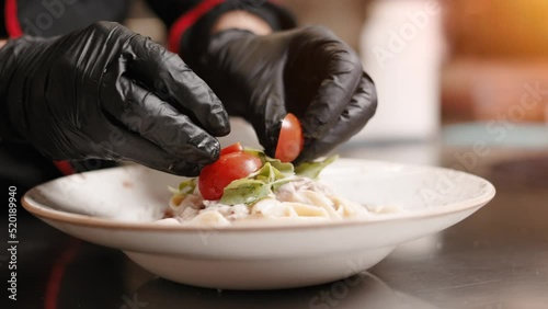 Close-up Professional Chef at Restaurant Kitchen Serves Dish Adding Final Ingredient by Hand. Chef adding tomato as decoration on pasta