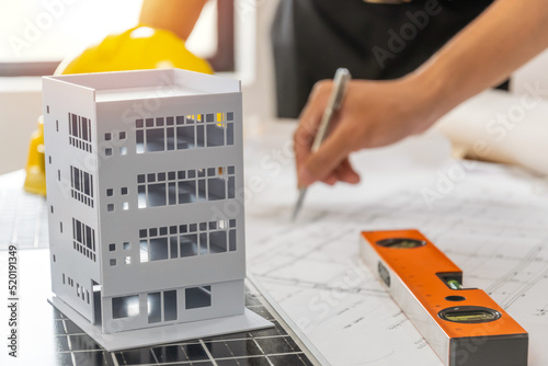 building model on workplace desk with architect or construction worker working with blueprint on desk in office center at construction site, industrial, construction contract and contractor concept photo