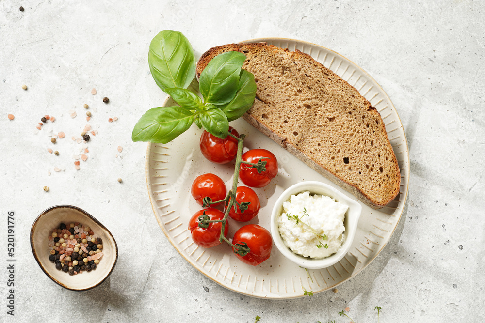 Roasted cherry tomatoes with branch, fresh cottage cheese, green basil and a slice of whole wheat bread on a plate on grey background