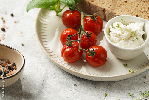 Roasted cherry tomatoes with branch, fresh cottage cheese, green basil and a slice of whole wheat bread on a plate on grey background