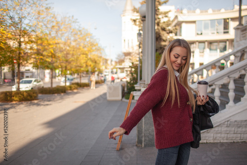 Style woman dressed casual sweater with t-shirt and jeans, walking outdoor with cup of coffee in the street of the city. Lifestyle portrait of young female model