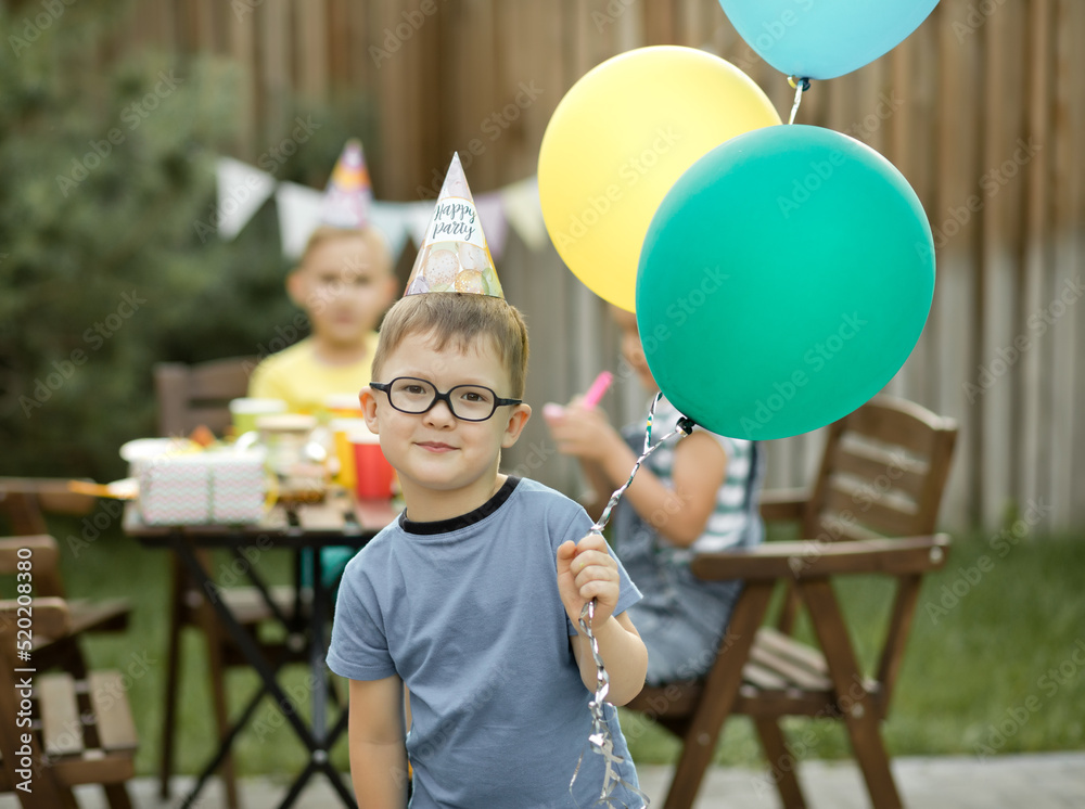 Cute funny four year old boy celebrating his birthday with family or friends in a backyard. Birthday party. Kid wearing party hat and holding balloon in a hand