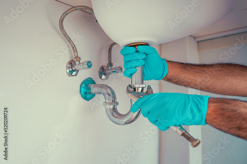 Plumber with blue gloves repairing sink siphon in kitchen. Plumber repairs and maintains chrome siphon under the washbasin. Refurbishment in the apartment. Elimination of leakage in the sink photo