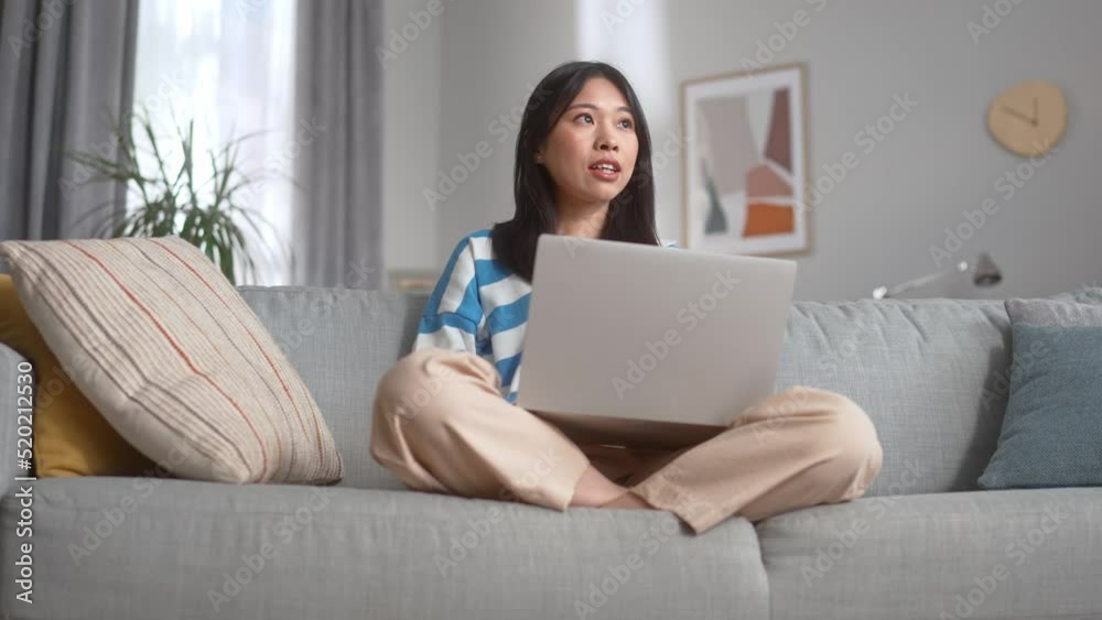 Happy woman resting on comfortable couch and using wireless laptop in bright apartment. Smiling attractive chinese female looking aside while typing messages in keyboard. Happy woman resting on comfortable couch and using wireless laptop in bright apartment. Smiling attractive chinese female looking aside while typing messages in keyboard.
