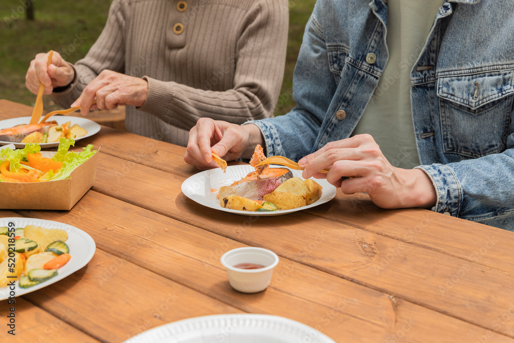 Salmon grilled fish with grilled vegetables in eco disposable tableware. Salat with pepper in eco disposable container. Two men eating salmon with grilled vegetables with wood fiber fork and knife.