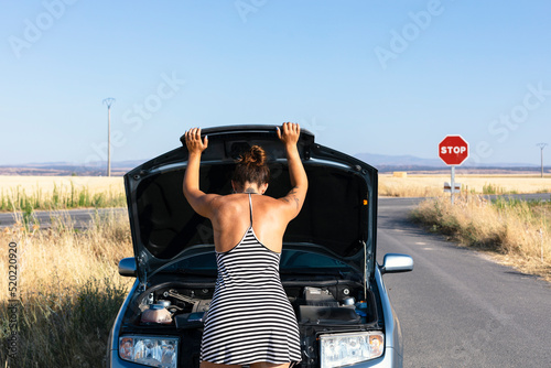 broken car engine on road. Young man lifting hood and looking at engine breakdown. Woman leaning on the hood of the car on a secondary road in the middle of a heat wave. Back portrait.