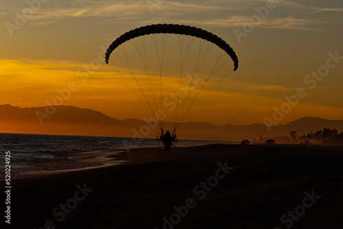 person in a motorised paraglider making a gliding flight on the shore of the beach at sunset or sunrise with an orange sky unrecognisable people
