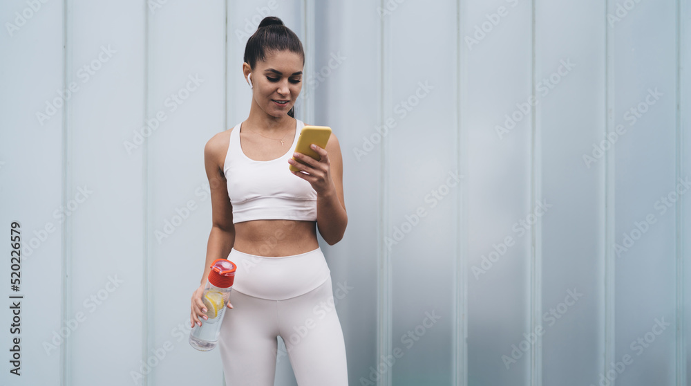 Portrait of young smiling female in wireless earphones standing with mobile phone device on copy space background. Attractive brunette woman with fit figure controls physical activity using mobile app Portrait of young smiling female in wireless earphones standing with mobile phone device on copy space background. Attractive brunette woman with fit figure controls physical activity using mobile app
