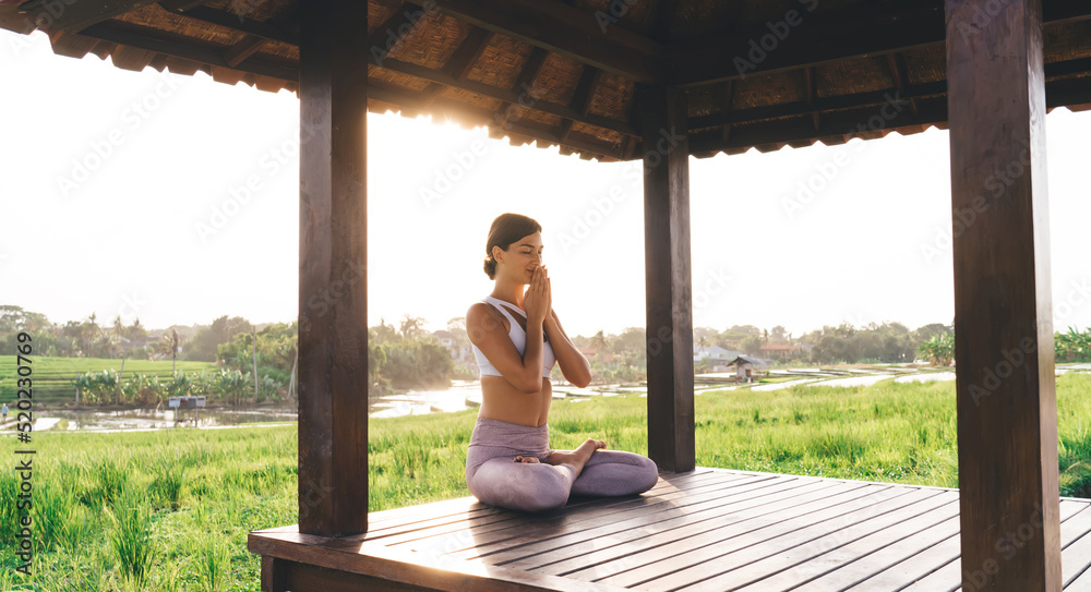 Side view of tranquil female in namaste praying near Indonesian rice fields and meditating for get mindfulness and enlightenment, calm woman in sportswear enjoying morning yoga practice in Thailand