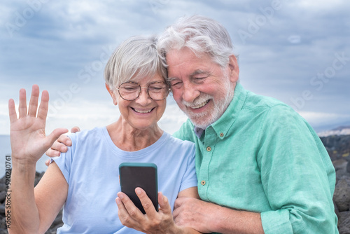 Beautiful happy senior couple sitting together on the cliff looking at smart phone. Cheerful elderly white-haired man and woman holding cellphone in video call