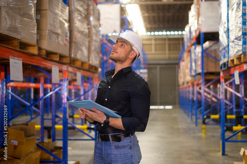 Fototapeta premium Young worker wearing helmet checking inventory and counting product on shelf in modern warehouse.