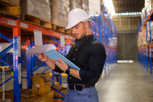 Young worker wearing helmet checking inventory and counting product on shelf in modern warehouse.