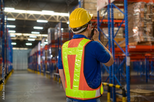 Young asian male worker wearing helmet using talki walki in modern warehouse.