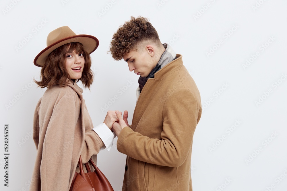 a cute couple stands on a light background dressed in coats and sweaters, a man stands against a woman and hugs her in front a cute couple stands on a light background dressed in coats and sweaters, a man stands against a woman and hugs her in front