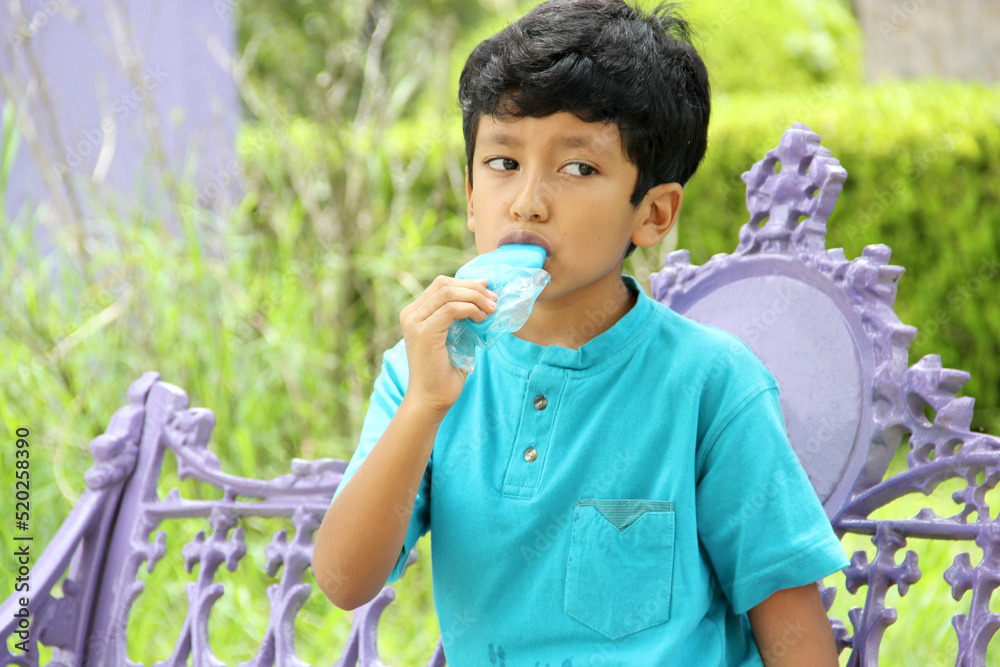 Fototapeta premium Poor Latino dark-haired boy with a blue t-shirt sitting on a park bench eating an ice pop because of the heat wave and cooling off in poverty by affecting his teeth with sweets and sugar