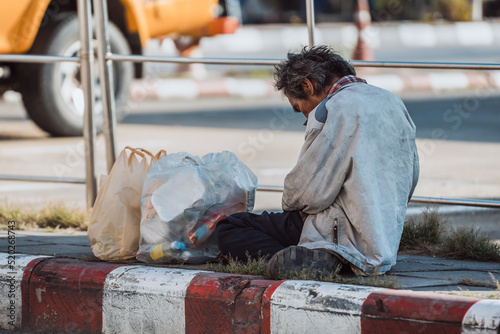 Homeless man sitting on roadside footpath in the big car background  and begging for help and money. Problems of big modern cities photo