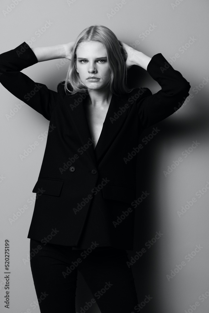 Pretty blonde female in black classic suit jacket touching hair with hand looks at camera posing isolated on grey studio background. Fashion concept