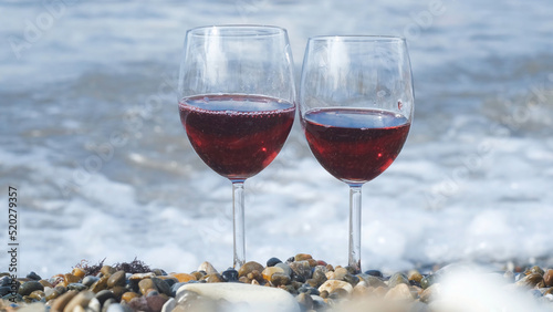 Two glasses of red wine on beach on background sea waves. Concept. Two glasses of wine are on beach for romantic date. Beautiful glasses of wine by sea