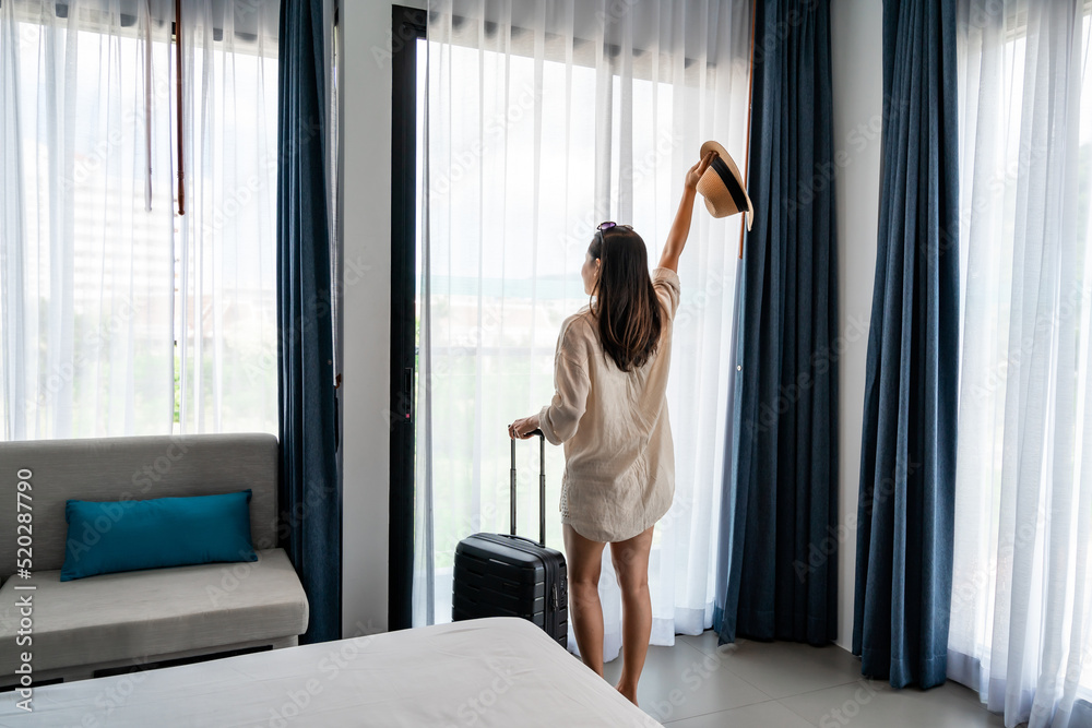 Young woman traveler with luggage looking at view in hotel room on summer vacation