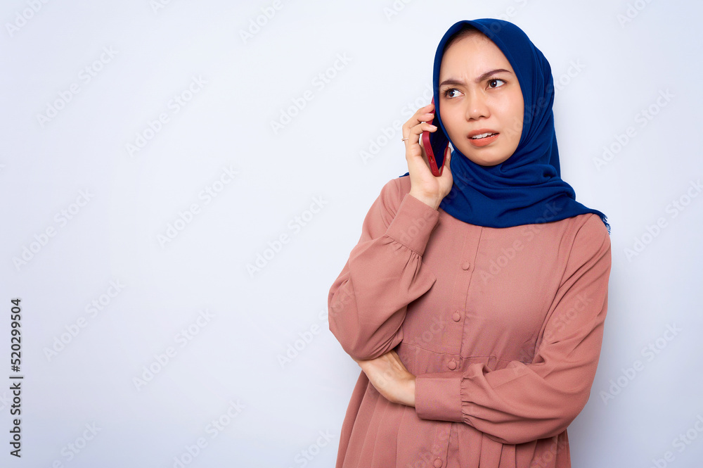 Confused young Asian muslim woman in pink shirt talking on mobile phone isolated over white background. People religious lifestyle concept