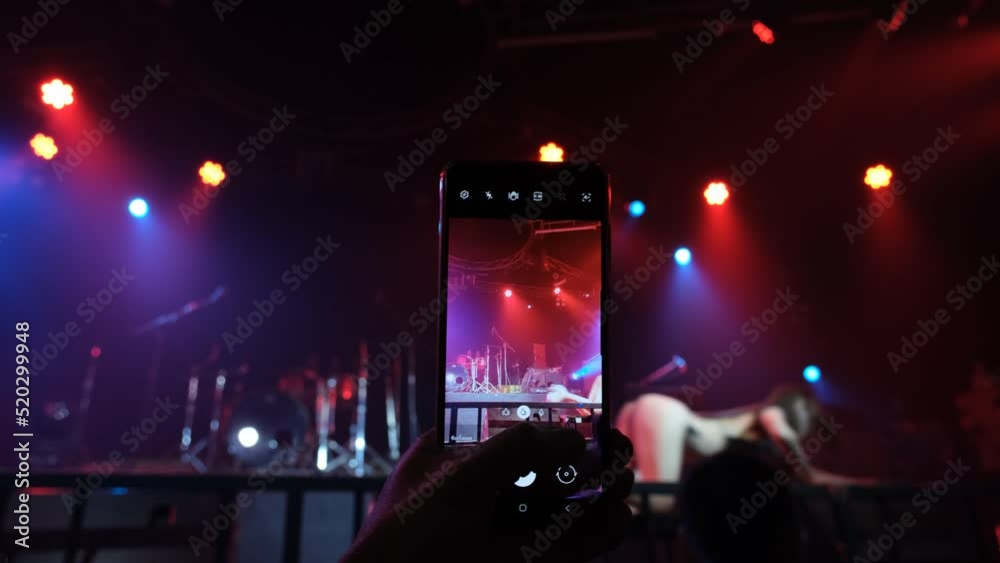 A man's hand holding a smartphone shoots a video of an Asian female model performing a sexy dance on stage.	