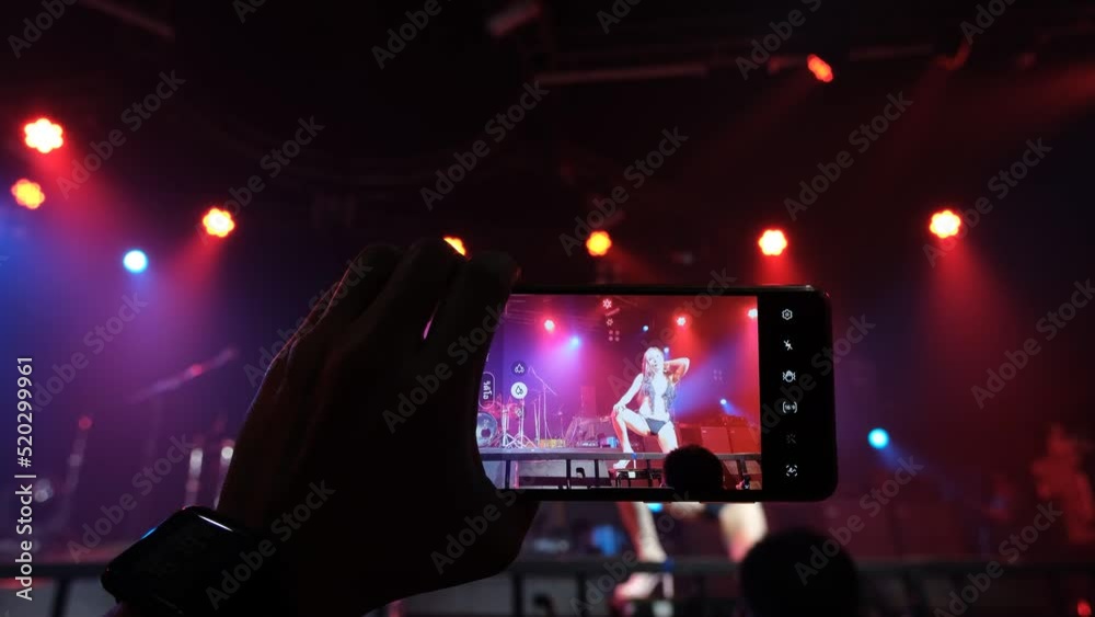 A man's hand holding a smartphone shoots a video of an Asian female model performing a sexy dance on stage.	