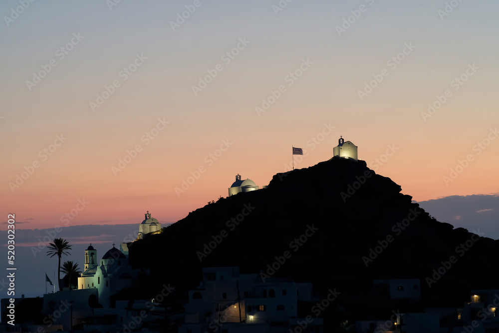 View of three illuminated small picturesque chapels, the Greek flag and an amazing colourful sunset in Ios Greece