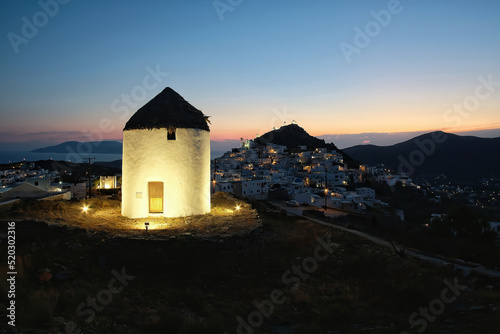 View of a beautiful illuminated white windmill, while the sun is setting dramatically behind the village of Ios in Greece 