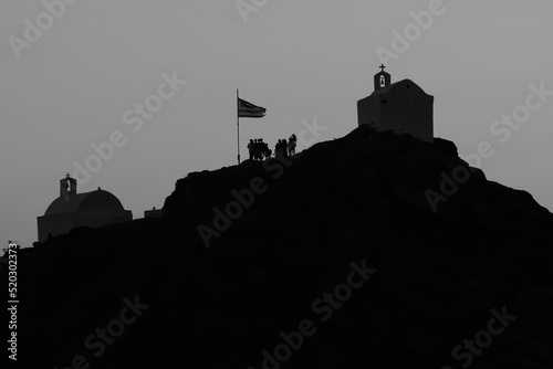Tourists exploring the picturesque small chapels up the hill  next to the Greek national flag   on the stunning island of  Ios Greece while the sun is setting in black and white