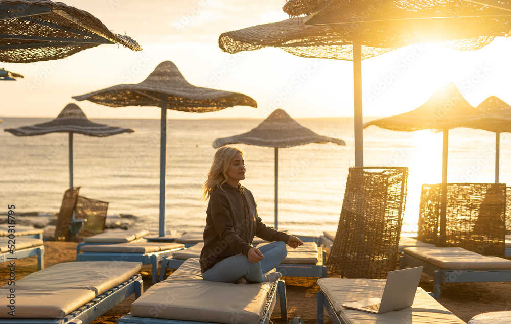 A calm woman sits on a sun lounger near the sea, gets distracted from working at the computer, meditates and removes negative emotions, combines remote work with rest.